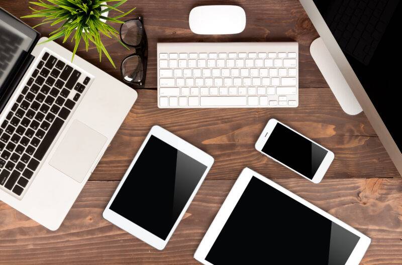 A MacBook, a pair of glass, a Mac mouse and keyboard, 2 iPads and an iPhone spread out on a desk. 